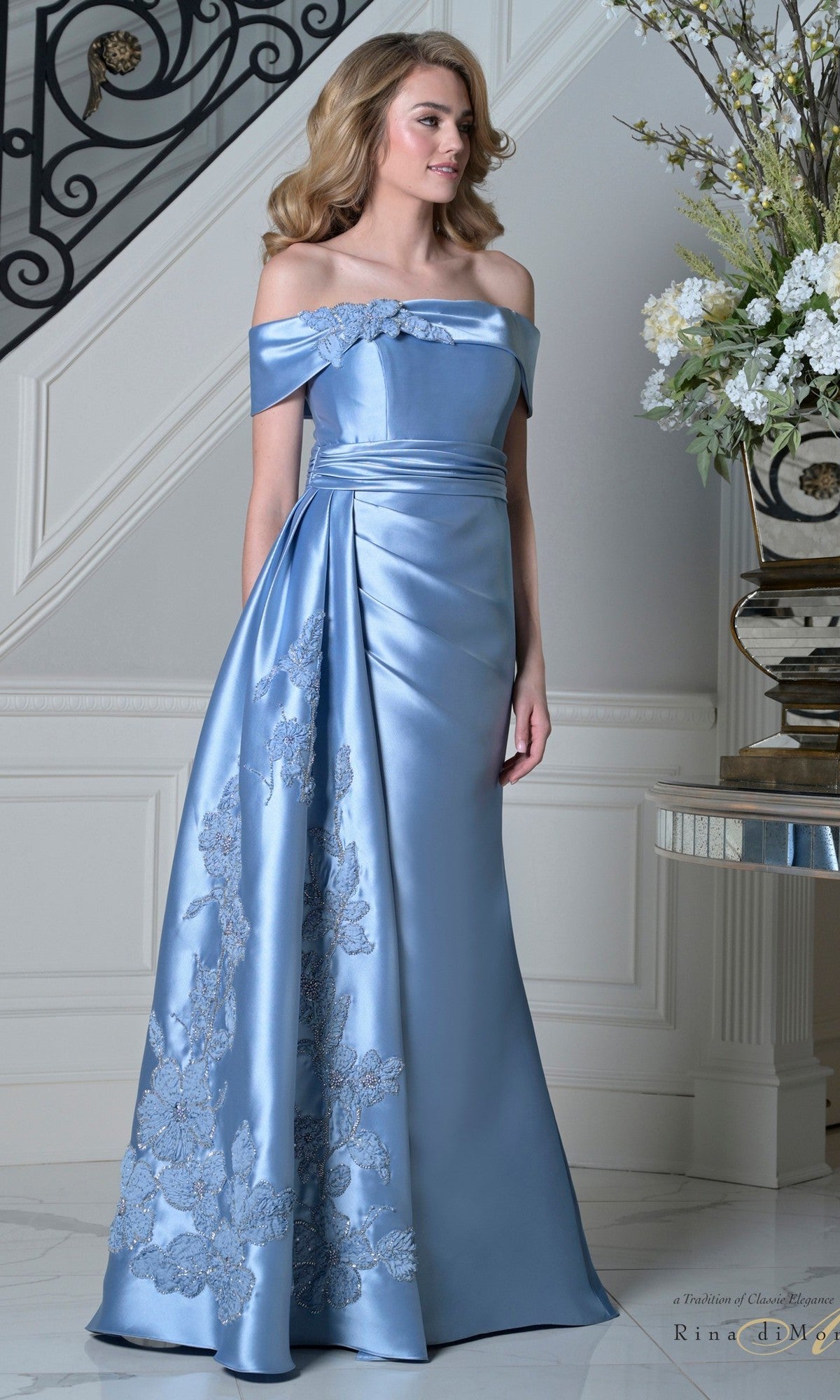Woman wearing the Long Formal Dress: Rina Di Montella RD3504 in sky blue satin with a ruched waistband and floral lace accents, standing indoors near a staircase and floral arrangement.